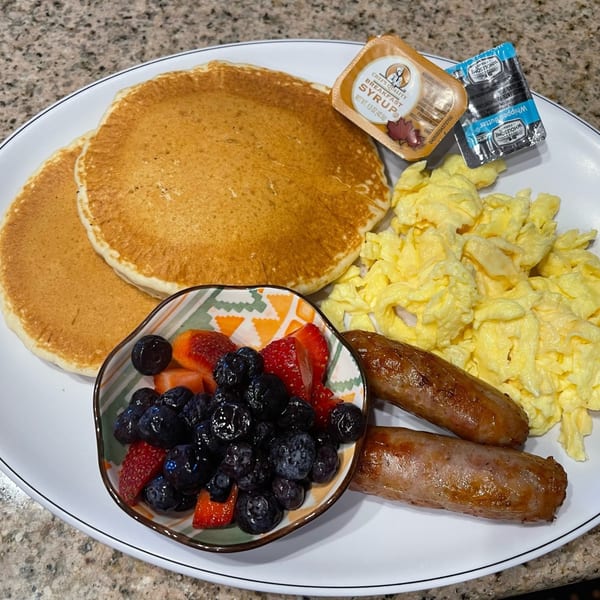 A breakfast plate with pancakes, scrambled eggs, sausage links, and a bowl of berries.