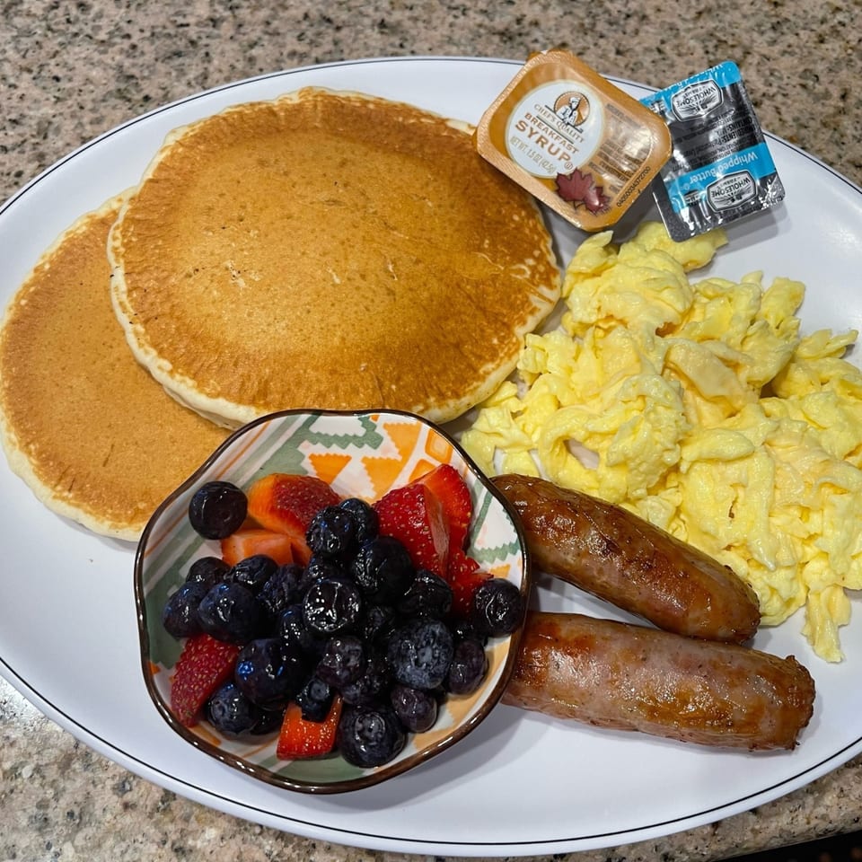 A breakfast plate with pancakes, scrambled eggs, sausage links, and a bowl of berries.