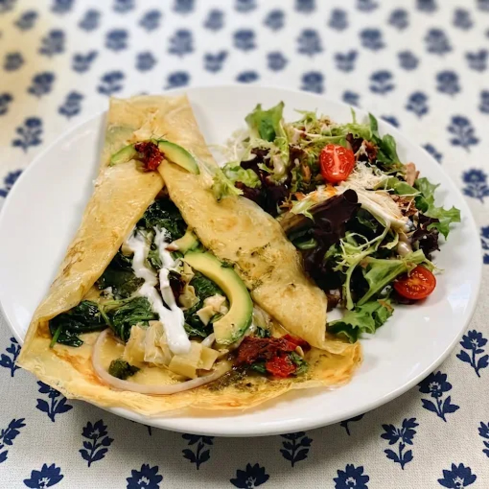Folded savory crepe filled with greens, avocado, mushrooms, and sun-dried tomatoes, served beside a mixed salad with cherry tomatoes.
