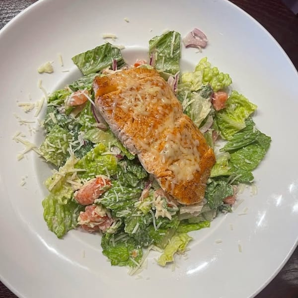 A grilled salmon fillet with melted cheese on a bed of Caesar salad with shredded parmesan.