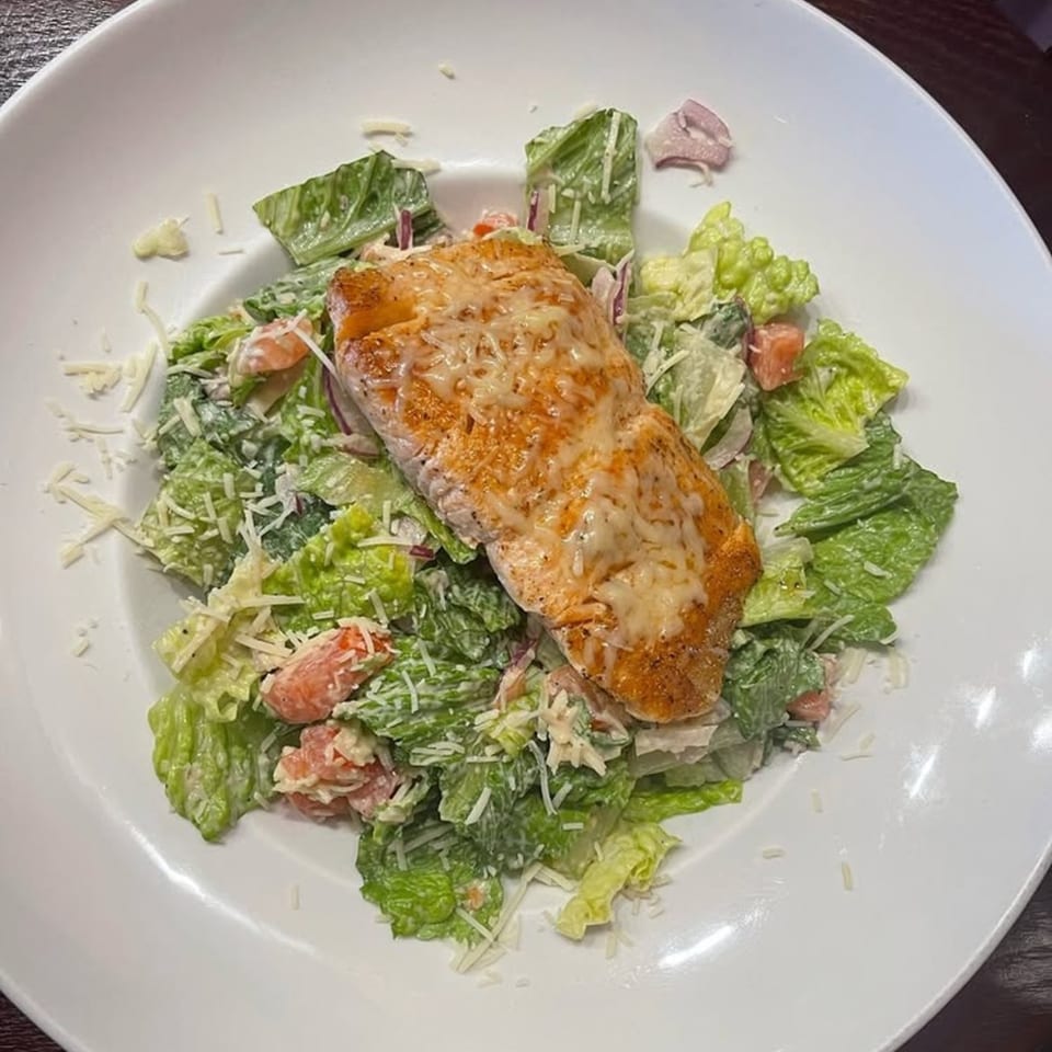 A grilled salmon fillet with melted cheese on a bed of Caesar salad with shredded parmesan.