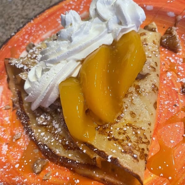 A golden crepe topped with whipped cream, sliced peaches, and powdered sugar on an orange plate.