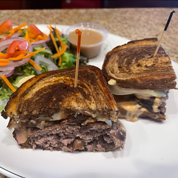 A grilled sandwich with meat, cheese, and onions, served with a side salad and dipping sauce.