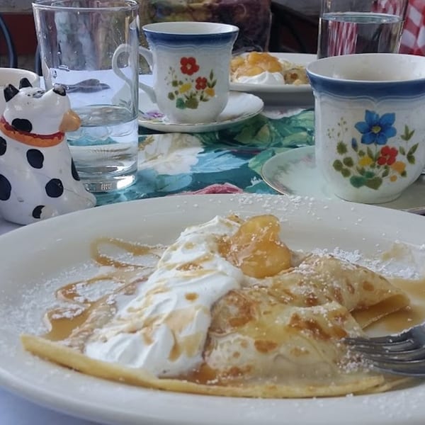 Crepes topped with whipped cream, fruit, powdered sugar, and syrup, with floral teacups on a table.