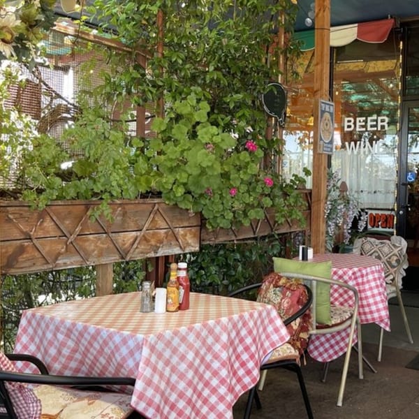 Outdoor patio seating at Crepe Town with red and white checkered tablecloths, lush greenery, and an "Open" sign.