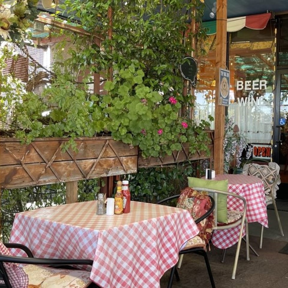 Outdoor patio seating at Crepe Town with red and white checkered tablecloths, lush greenery, and an "Open" sign.