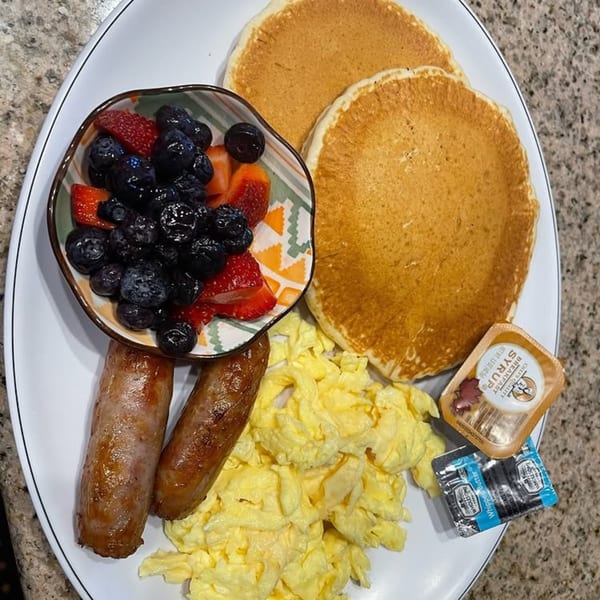 A breakfast plate with pancakes, scrambled eggs, sausage links, and a bowl of berries.