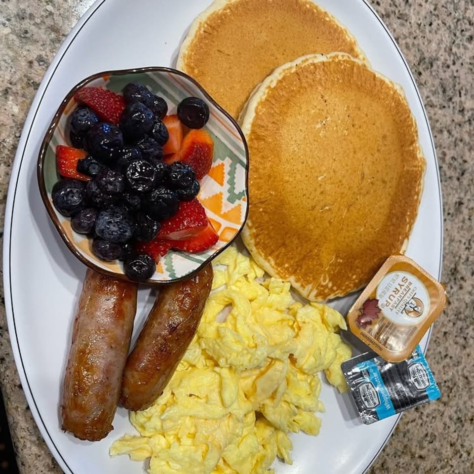 A breakfast plate with pancakes, scrambled eggs, sausage links, and a bowl of berries.