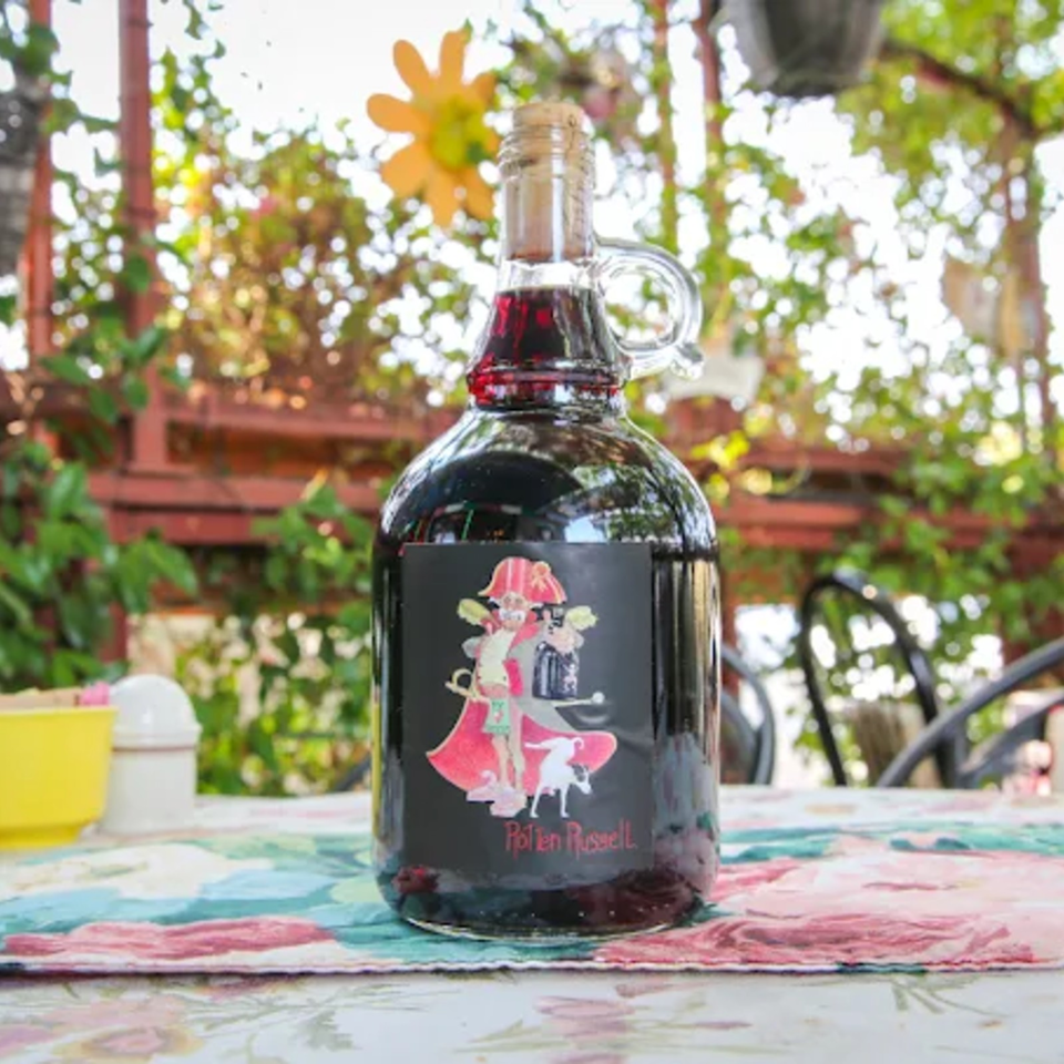 Glass bottle of dark red Apéritif Russell liquor on a colorful outdoor table, surrounded by plants and sunlight.