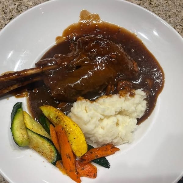 Braised lamb shank with mashed potatoes and roasted vegetables on a white plate.