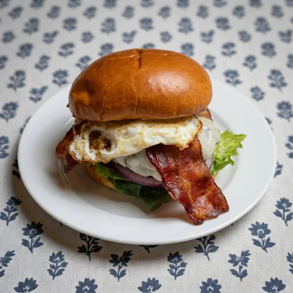Bacon cheeseburger with a fried egg, lettuce, and red onion on a white plate over patterned fabric.