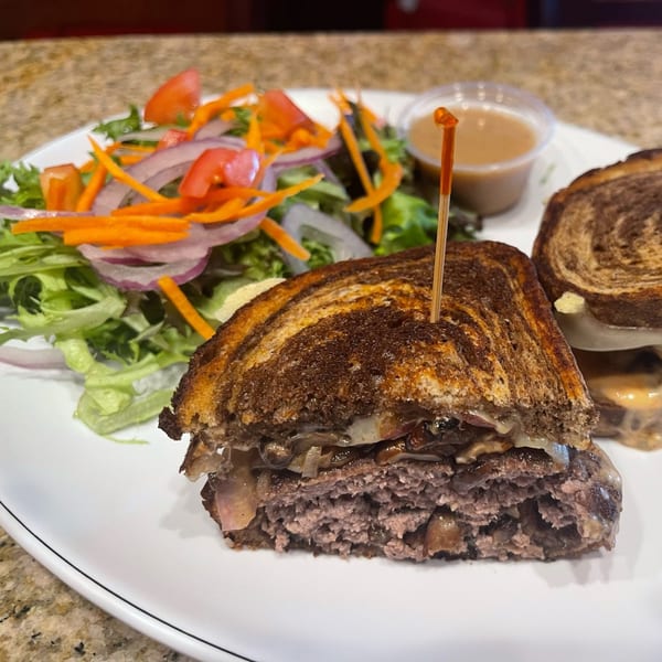 A grilled burger sandwich with mushrooms and cheese, served with a side salad and dipping sauce.
