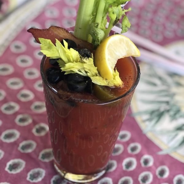 Bloody Mary cocktail garnished with bacon, celery, olives, and a lemon wedge on a patterned tablecloth.