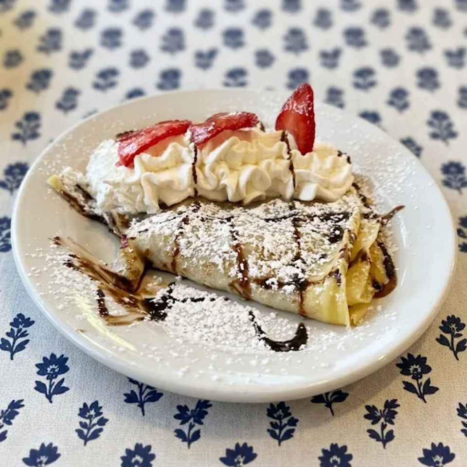 Crepe topped with whipped cream, strawberries, powdered sugar, and chocolate sauce on a white plate.